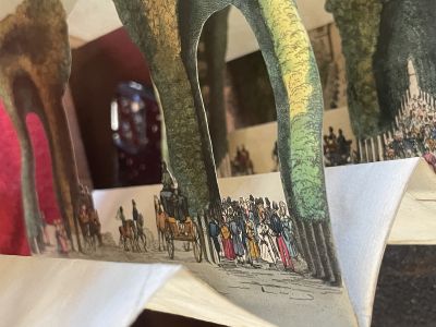 A view of a cutout page showing crowds of people in colorful outfits beneath the arching trees. At the bottom of the photo, note the partially folded paper strips that form the sides (or bellows) of the tunnel book.